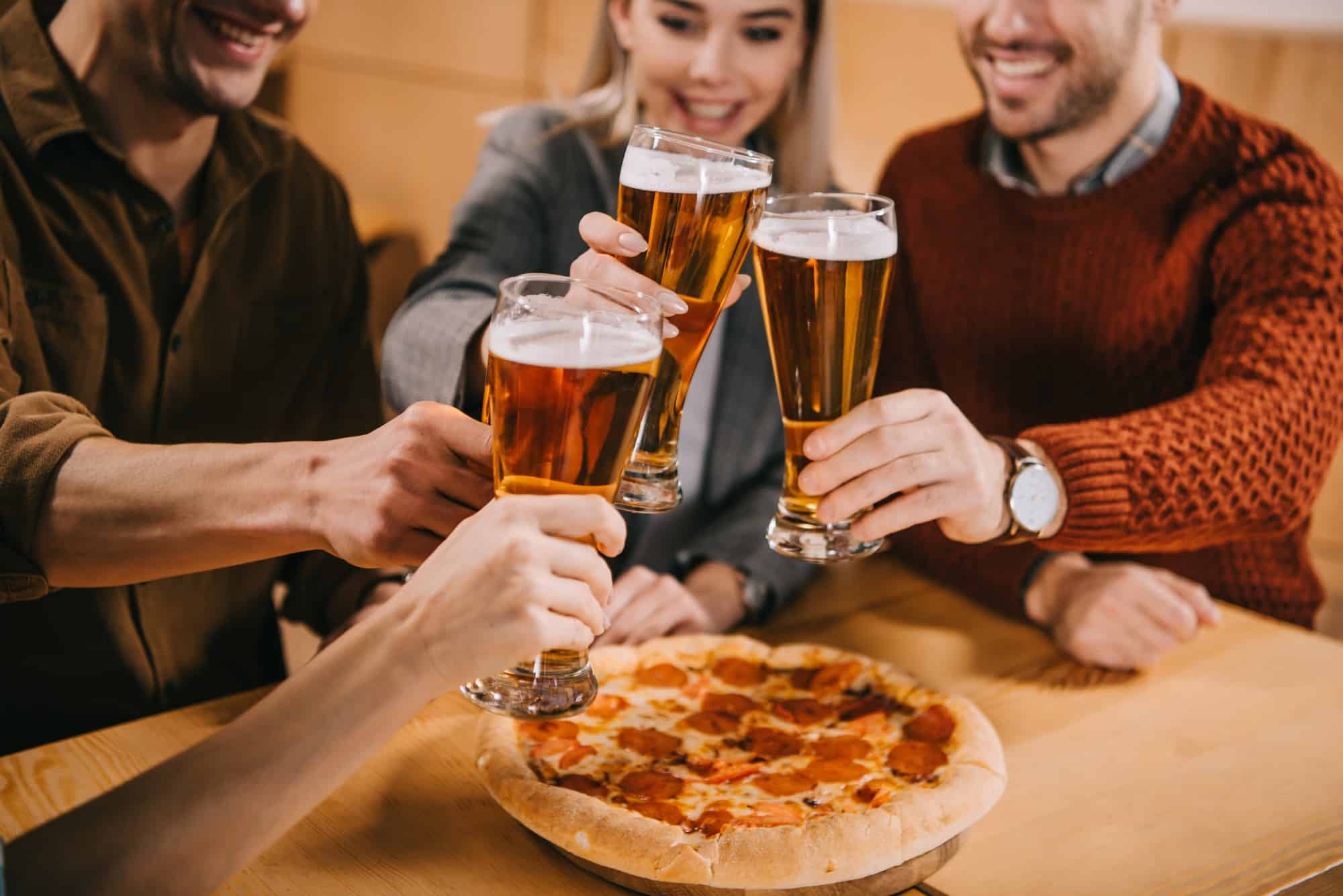cropped view of friends clinking beer near pizza in bar